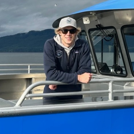 IMG_7931 Person on a boat deck with sunglasses, cap, and mountains in the background.